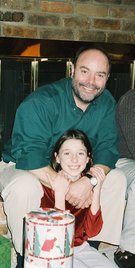 Dave seated in front of the fireplace, with Kimberly sitting between his knees, and a festively wrapped Christmas package