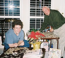 Grandpa leans over a card table where a puzzle is in the midst of assembly, while Kevin leans over the coffee table where a smaller number of puzzle pieces are arranged.