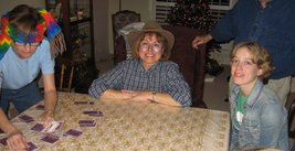 Nancy with a colorful scarf wrapped around her head deals the cards while Kathy (in a fedora) and Jocelyn sit at the table